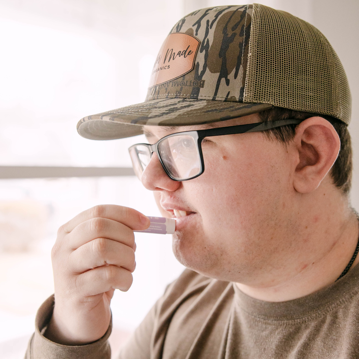 A man in profile wearing a camouflage trucker hat with a tan leather patch reading 'Taylor Made Organics' and black-framed glasses, applying lip balm to his lips.