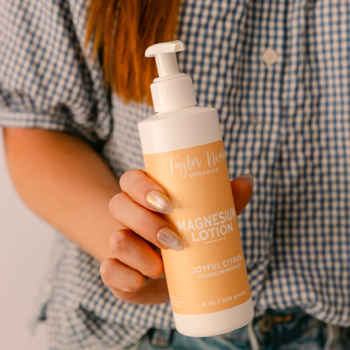 Hand holding Taylor Made Organics Joyful Citrus Magnesium Lotion with orange and grapefruit in an 8-ounce pump bottle.