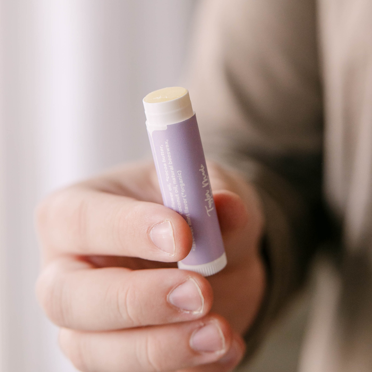 Hand holding an open tube of Taylor Made Organics Lavender Mint Lip Balm with a purple label.