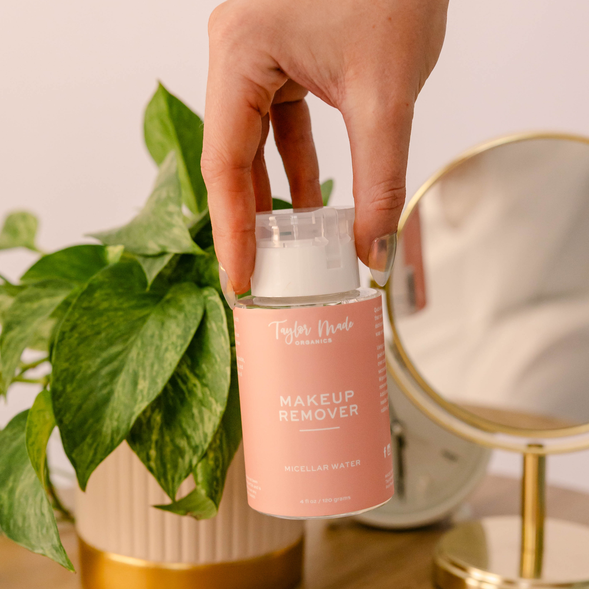 Hand holding Taylor Made Organics Micellar Makeup Remover beside a green plant and vanity mirror.