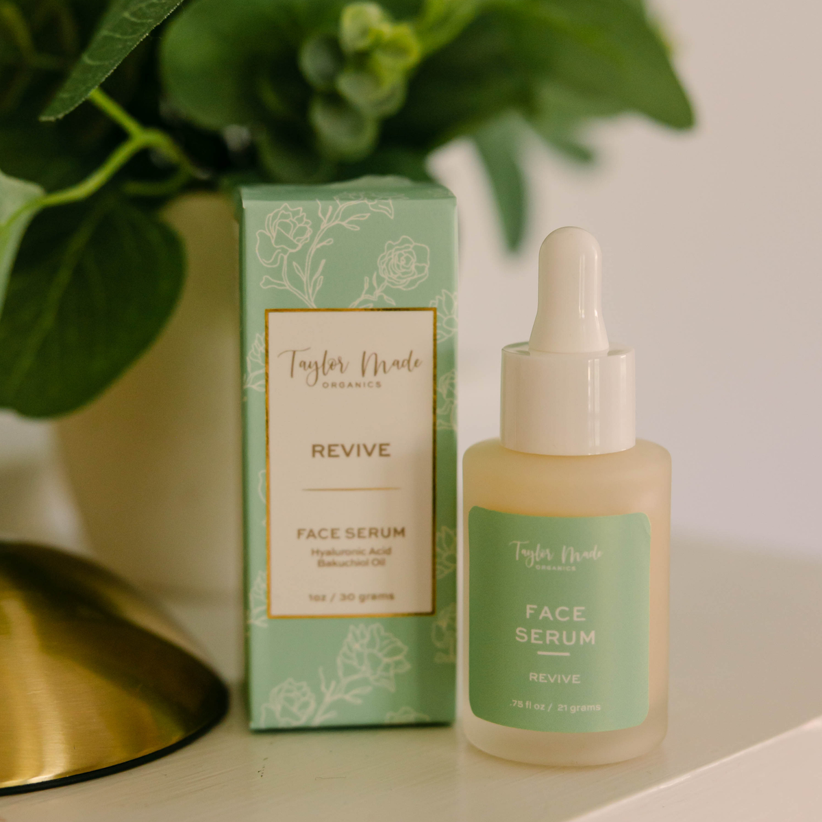 Taylor Made Organics Revive Face Serum dropper bottle and box displayed beside a green houseplant.