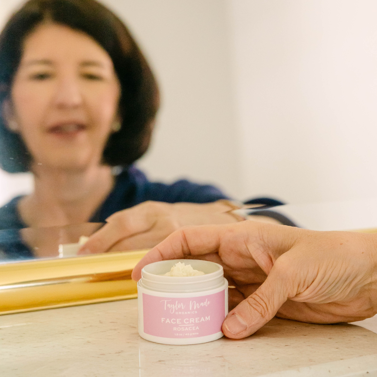 Woman holding an open jar of Taylor Made Organics Rosacea Face Cream in front of a bathroom mirror.
