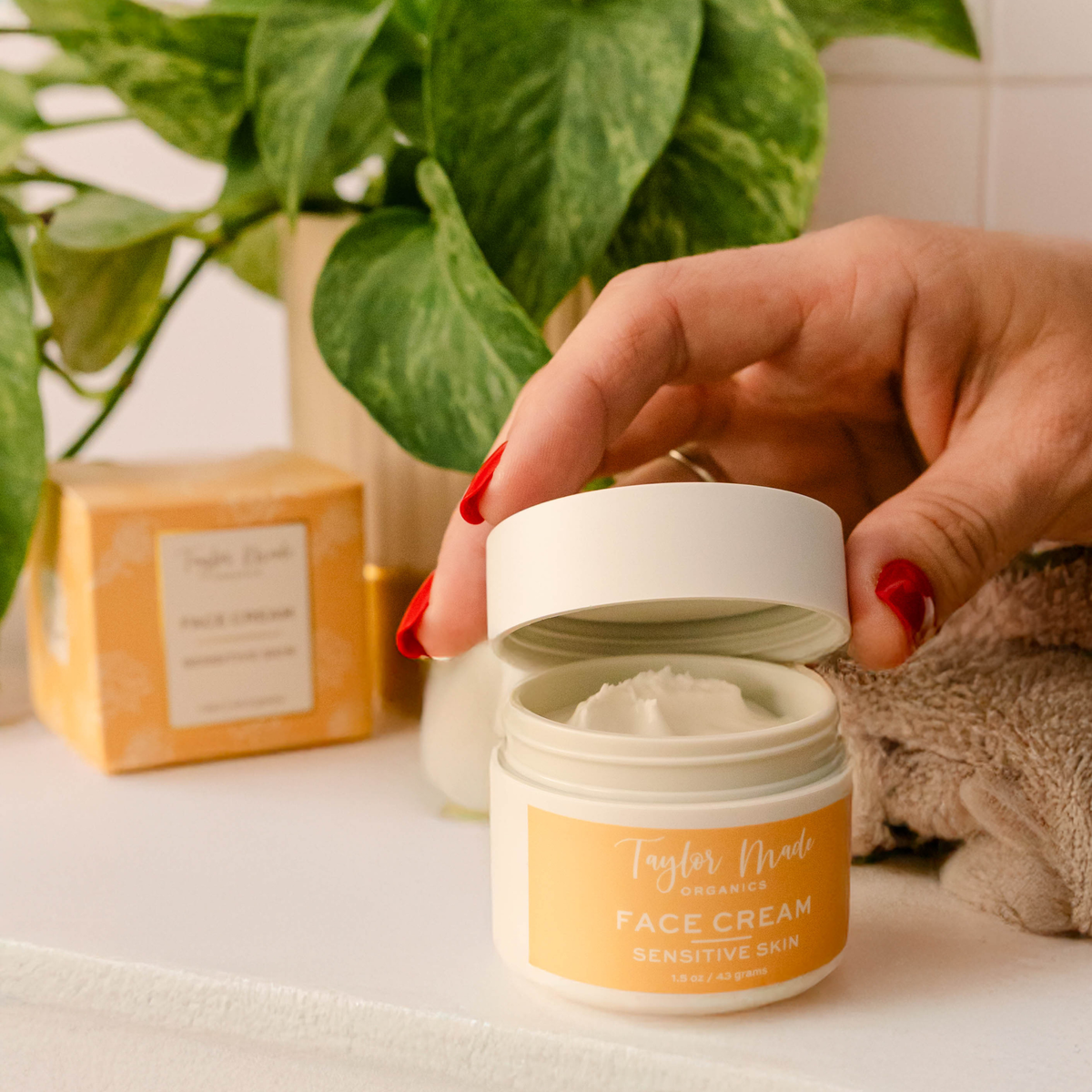 Hand opening a jar of Taylor Made Organics Sensitive Skin Face Cream beside a green houseplant.